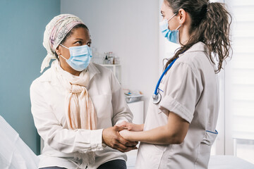 Young female doctor in medical uniform and stethoscope wearing face mask speaking and holding hands of African American mature woman patient during appointment in clinic