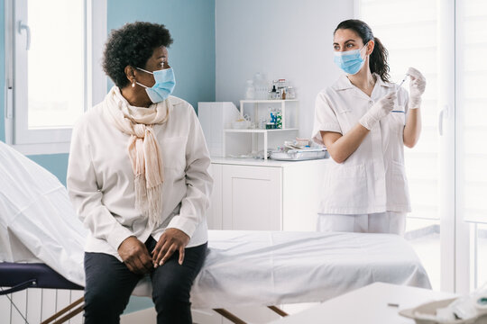 Female doctor in latex gloves and face shield filling in syringe from bottle with vaccine preparing to vaccinate unrecognizable mature African American woman patient in clinic during coronavirus outbreak