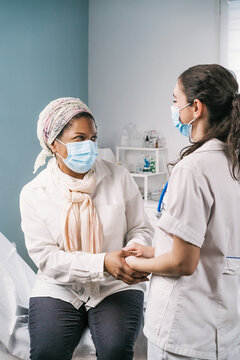 Young female doctor in medical uniform and stethoscope wearing face mask speaking and holding hands of African American mature woman patient during appointment in clinic