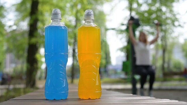 
Two blue and orange bottles of isotonic drink and a woman doing sports on a simulator outdoor, summer day.