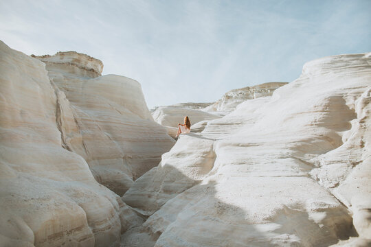 Back view anonymous female in white sundress strolling along narrow passage formed by light curvy rocks on sunny weather in Sarakiniko Greece