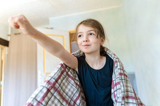 Happy little girl in casual shirt with soft blanket on shoulders outstretching arm forward imagining flight and looking away in light bedroom