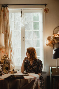 Young mindful female looking away at table with books against window in house on sunny day