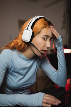 Side view of worried female gamer in headphones playing video game while sitting at table with keyboard