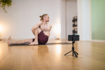 Soft focus of smartphone on tripod recording yoga session of coach practicing Marichyasana on sports mat in studio