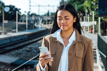 Cheerful Asian female traveler standing on platform at railway station and messaging on mobile phone