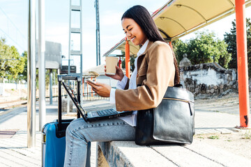 Side view of happy Asian female freelancer sitting on bench with laptop and takeaway coffee and browsing smartphone while waiting for train at railway station and working remotely