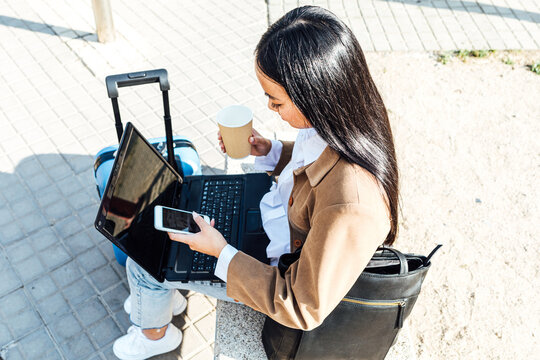 From Above Side View Of Asian Female Freelancer Sitting On Bench With Laptop And Takeaway Coffee And Browsing Smartphone While Waiting For Train At Railway Station And Working Remotely