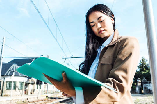 From Below Focused Asian Female Entrepreneur Reading Documents While At Railway Station And Waiting For Departure