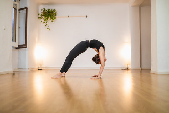 Side view of unrecognizable flexible female in sportswear standing in Urdhva Dhanurasana pose while practicing yoga on shiny floor