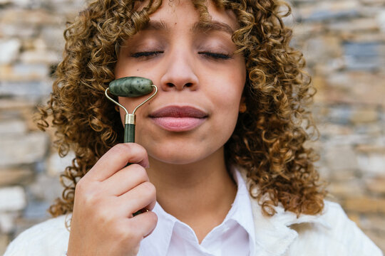 Dreamy African American female using jade roller and doing facial massage while standing with closed eyes in park