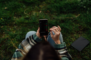 From above of crop anonymous female dealer touching screen of cellphone with graphs while sitting on meadow