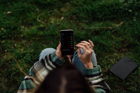 From above of crop anonymous female dealer touching screen of cellphone with graphs while sitting on meadow