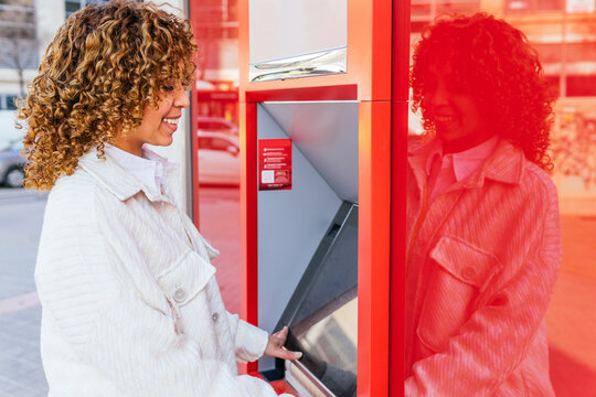 Side view of smiling African American female using ATM terminal and withdrawing cash while standing on city street