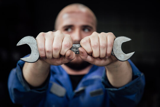 Faceless male mechanic in blue overall covering face with metal spanner in hands looking away on black background