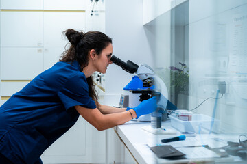 Side view of female vet in blue uniform looking through microscope while working in lab