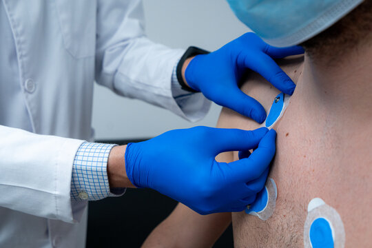 Crop medic applying electrodes on chest of unrecognizable male patient in mask for ECG test