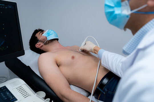 Side view of cropped unrecognizable male medic using modern ultrasound equipment and examining heart of male patient in mask lying on medical table during diagnostic in hospital