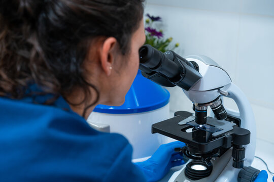 Back view of female vet in blue uniform looking through microscope while working in lab