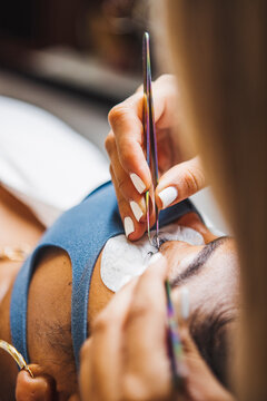 Crop unrecognizable cosmetologist with tweezers applying fake eyelashes for extension on eye of ethnic client with face protective mask in salon during coronavirus pandemic