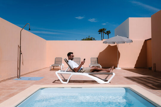 Side view of male freelancer lying on lounger at poolside and surfing Internet on mobile phone during telework in summer on sunny day