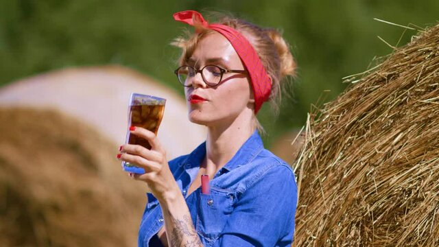 Blonde girl drinks a soft drink on a sultry day on a countryside field. Sultry summer day and a girl on the background of haystacks in the field during the harvest. Coca Cola with ice