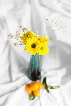 Bright blossoming chrysanthemums with pleasant aroma and sprigs in vase on crumpled white textile with fresh kumquats