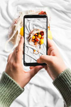 Overhead view of crop unrecognizable person touching screen on mobile phone while taking photo of fruits in zero waste bag
