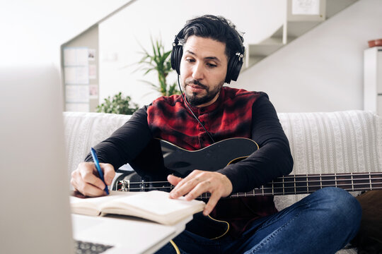 Adult male guitarist in headphones with electric guitar taking notes in notebook while composing music against netbook on sofa at home