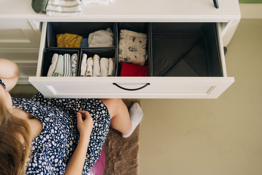 From above of crop unrecognizable expectant female sitting against chest of drawers with row of wear in house room