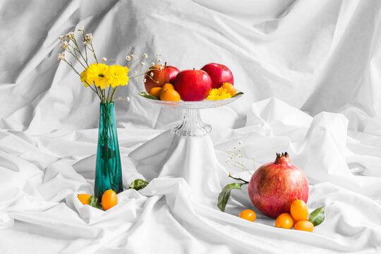 Tasty pomegranate with red apples and kumquats near vase with blooming chrysanthemum on white creased fabric
