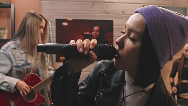 Slowmo closeup of young attractive rock woman in cool leather jacket and beanie hat singing in microphone while her female colleagues from band playing musical instruments in retro-style studio