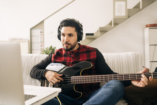 Adult male musician in headphones playing bass guitar against netbook on sofa in living room