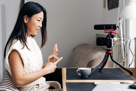 Smiling ethnic female blogger with cup of hot drink talking while recording video on photo camera at home