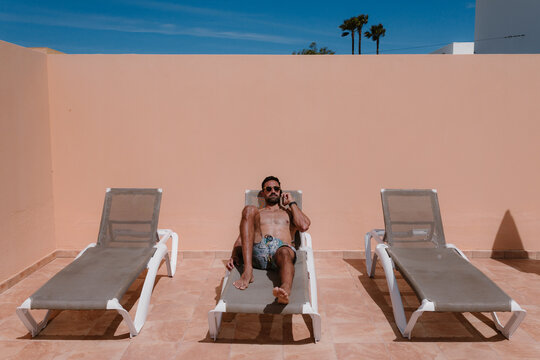 Focused male freelancer lying on lounger and sunbathing while discussing project on smartphone and working remotely in summer