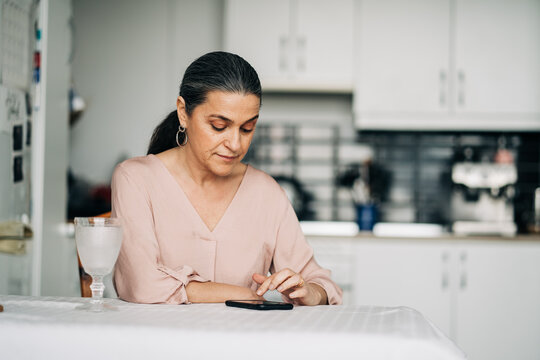 Concentrated Middle Aged Female Surfing Cellphone While Sitting At Table In Modern Kitchen With Kitchenware At Home