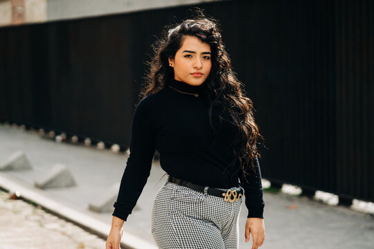 Plus size female wearing trendy cloths standing against black wall in city and looking at camera