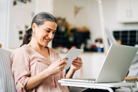 Cheerful middle aged female sitting on couch near modern netbook while looking at photo in modern apartment with kitchen on blurred background