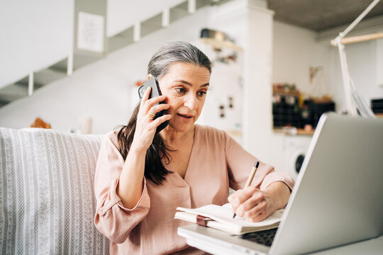 Concentrated middle aged female having phone call and taking notes in notepad while looking at screen of laptop in light room at home