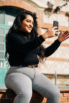 Cheerful plus size female in trendy clothes sitting on fence and taking self shot on smartphone on sunny day in city