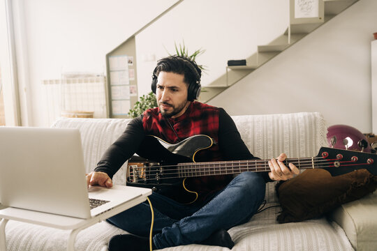 Adult male musician in headphones playing bass guitar against netbook on sofa in living room