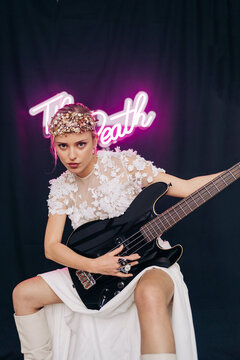Energetic rebellious young woman in elegant white bridal dress and wreath with guitar in hand making horn gesture in studio with neon inscription