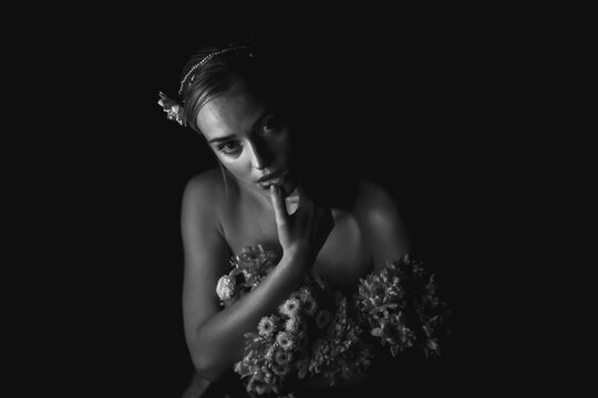 High angle black and white of charming young bare shouldered female with bunch of flowers and floral adornment on head touching lips and looking at camera in dark studio