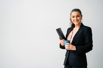 Positive middle aged female entrepreneur with ponytail wearing formal suit looking at camera while standing with folder and cup of drink on white background