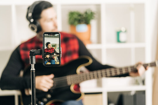 Tripod with cellphone screen representing photography of male musician in headset playing bass guitar in house