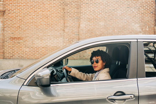 Side view of African American female driver in stylish sunglasses and fashionable outfit smiling while driving modern automobile on the street