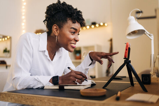 Side view low angle of African American female writing in notebook while watching online tutorial via mobile phone at home