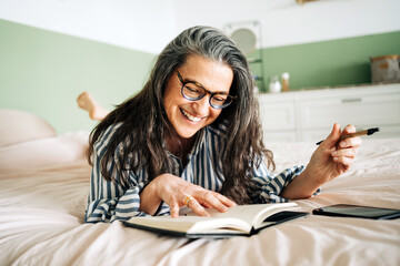 Middle aged cheerful female in striped shirt smiling and reading notebook on bed with smartphone