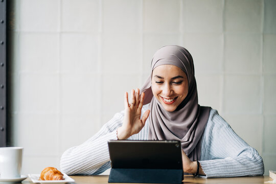 Content Muslim female in hijab waving hand and talking on video chat via tablet while sitting at table in cafe