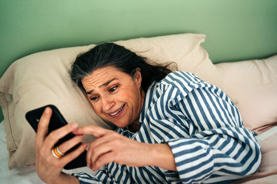 From above of amazed middle aged Hispanic female with long dark hair in striped sleepwear smiling happily while reading good news on smartphone lying in bed